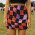 Honey Dazed mini pencil skirt featuring a bold wavy checkerboard pattern in black, orange, purple, and pink. Made from soft eco-polyester with a curve-hugging stretch fit and a flattering high-waist silhouette. The print has a slightly glossy finish, giving it a sleek and modern look. Designed for festival fits, streetwear moments, and retro rave energy. A vivid, sculptural mini skirt that pairs easily with crop tops and confident energy.